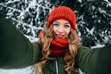 Winter happy selfie. Young beautiful happy girl in red winter knitted hat and scarf takes selfie in winter forest