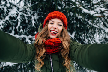 Winter selfie. Young happy girl in red knitted hat and scarf takes selfie and play the ape in winter cold day. Girl takes winter selfie