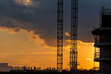 Obraz premium Construction workers silhouettes, cranes and building construction site against sunset sky.