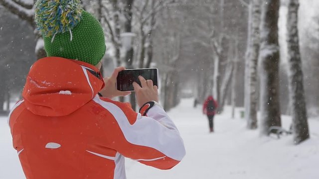 Slow motion of woman photographing winter park with snowfall on her phone
