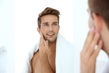Young man standing in front of mirror and touching his face