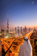 Naklejka premium Arabian man watching night cityscape of Dubai with modern futuristic architecture in United Arab Emirates