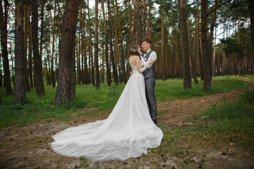 Wedding couple outdoor, beautiful bride in white wedding dress with long plume and elegant groom in grey suit with vest hugging in forest