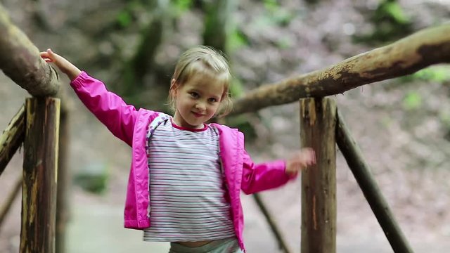 Funny little girl in pink jacket on the nature. Little girl stands on a wooden bridge and smiles