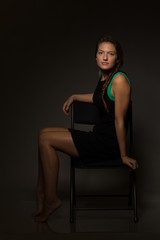 Fototapeta premium Young caucasian woman in a short black dress sits on a chair under moody light