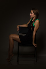 Fototapeta na wymiar Young caucasian woman in a short black dress sits on a chair under moody light