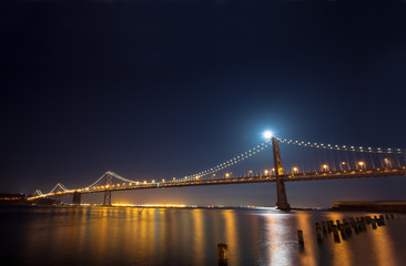 Fototapeta premium San Francisco Bay Bridge to Treasure Island at night. Panoramic view with a full moon shinning behind the bridge.