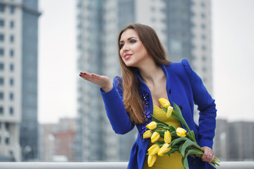 Young attractive girl in a blue coat and yellow dress holding a bouquet of yellow tulips. Spring is coming to town. She sends an air kiss