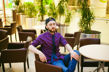 Portrait of a young man with a beard, dressed in jeans and a plaid shirt, tie and cap, sitting on a chair at the coffee shop in the mall