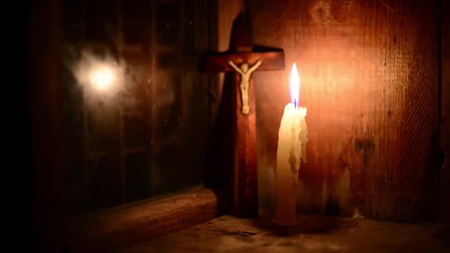 A candle burns on the window. Outside night. The cross with a crucifix standing behind. (Focus on candle). Candle flame flickers. Wooden windows frame in a wooden house in the mountains.