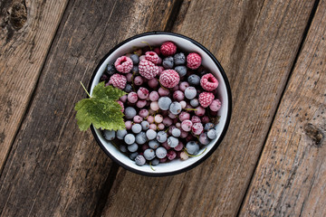 frozen berries, black currant, red currant, raspberry, blueberry in enamelled bowl decorated by currant leaf on wooden table in rustic style,  top view.