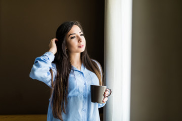 Attractive sexy woman with neat body is holding a cup with hot tea or coffee and looking at the sunrise standing near the window