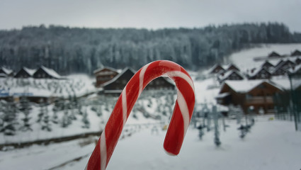 Close up of a red and white candy cane on a snowy background with snow covered houses and trees. Sweet caramel lollipop symbolizing the winter holidays.