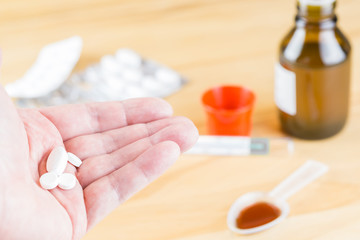 Eine Hand mit Tabletten im Hintergrund sieht man einen Löffel Hustensaft, eine Flasche mit...