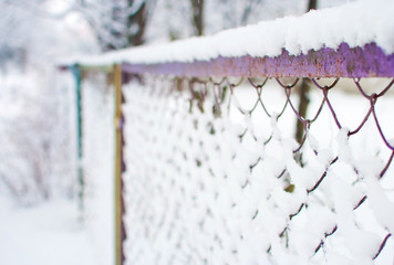 Closeup of a purple mesh fence covered in a thick layer of white fresh fluffy snow against blurred trees on the background. Cold winter day in January