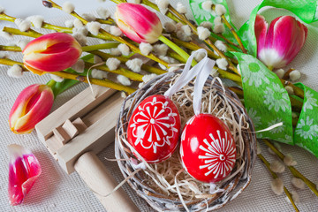 Traditional Czech easter decoration - regional wooden ratchet instrument with painted red eggs in wicker nest with pussycats and tulips flower. Spring easter holiday arrangement.