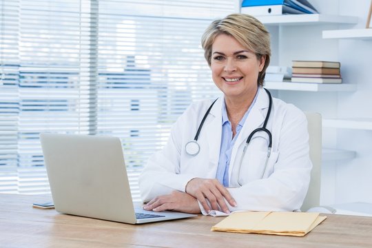 Portrait Of Smiling Female Doctor Sitting At Desk With Laptop