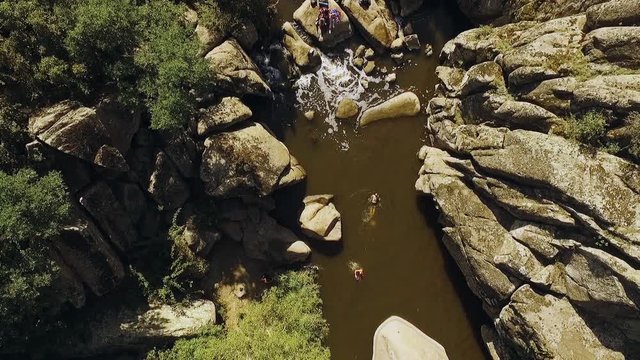 Friends swimming in river near small waterfall with husky dog playing frisbie. Panoramic shot from drone in slowmotion