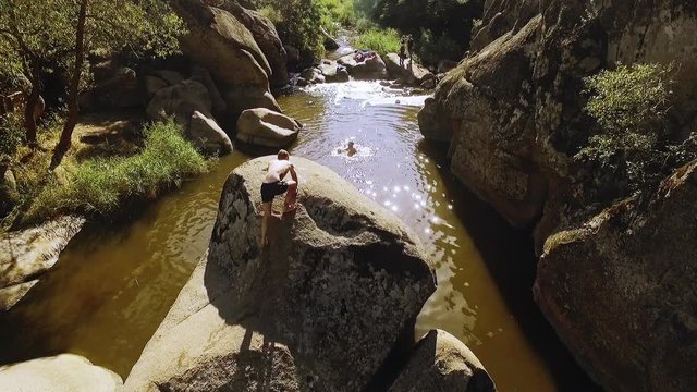 Male climbing big stone in river raising tattooed hands in front of friends. Panoramic shot in slowmotion from drone