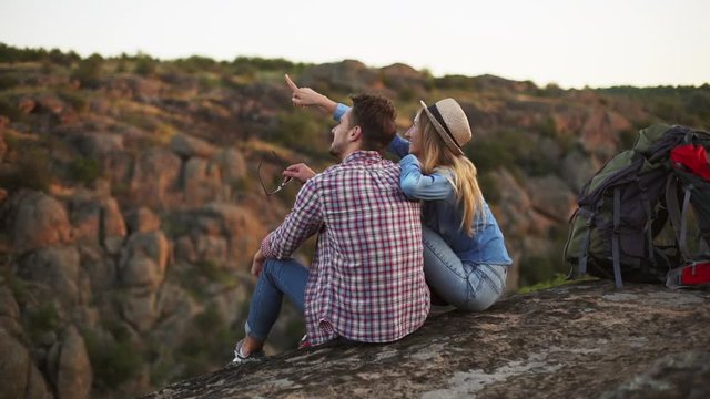 Caucasian young pair sitting on rock and smiling. She points her hand he holds sunglasse. Shot slowmotion from drone
