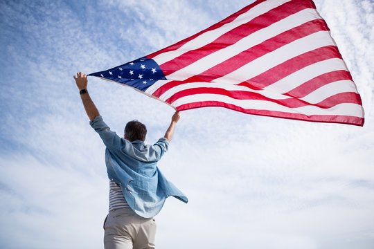 Rear view of man holding american flag on beach - Powered by Adobe