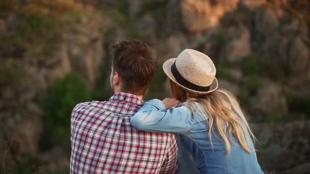 Young male in plaid shirt sitting with girlfriend in jeans shirt and hat. She puts hand on his shoulder. Back view in slowmotion