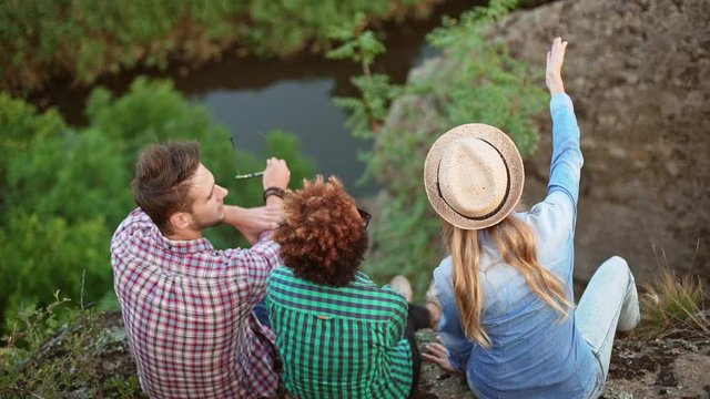 Male and female friends in shirts sitting on cliff above river admiring discussing view and smiling. Back view in slowmotion