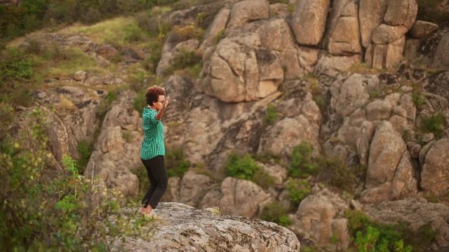 Young dark skinned girl with afro haircut wear green shirt and glasses dancing outdoors on rock among rocky landscape smiling waving hands in slowmotion