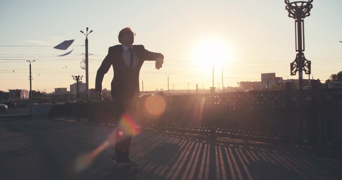 Young businessman riding skateboard on street at sunset and tossing up papers