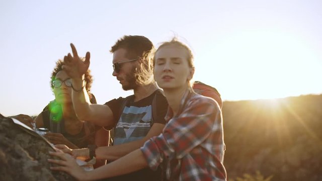 Three friends trying to find right direction using map and compass near big stone with sun on background. In slowmotion
