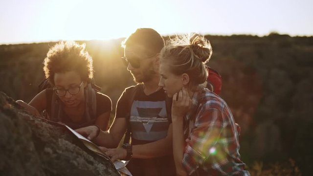 Three hikers with map discussing right direction near big rock outdoors in slowmotion