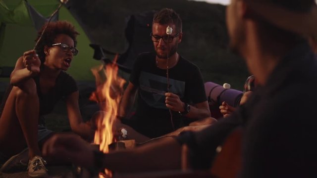 African American female in glasses singing song while her friends eating fried marshmallowand playing guitar in camp at night. Close up slowmotion