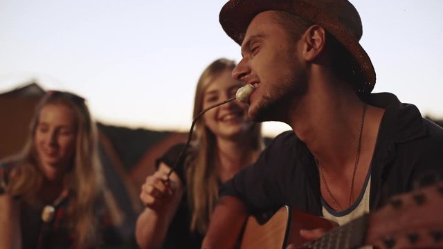 Caucasian male with bristle and hat playing guitar and eating fried marshmallow with two blonde females listening laughing smiling in slowmotion