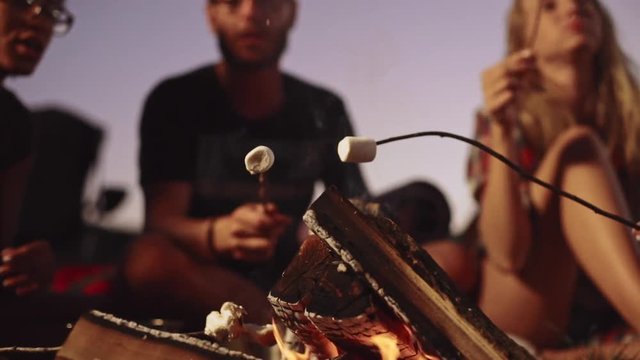 Group of friends frying marshmallow sitting by fire talking smiling.Close up view in slowmotion