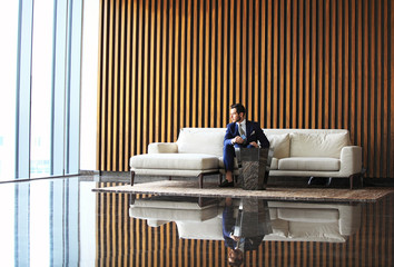 Happy young businessman sitting relaxed on sofa at hotel lobby using smartphon, waiting for someone.