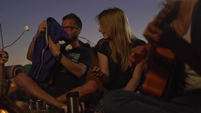 Caucasian male with beard and glasses covers blonde beautiful female with sport jacket while their friends sitting by fire playing guitar smiling talking. In slowmotion