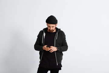 Bearded young man dressed in all black and navy casual clothes looks into his smartphone isolated on white