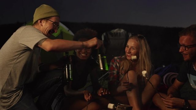 Caucasian male in green hat with glasses brings bottled beer to their friends in camping giving high five smiling laughing. In slowmotion
