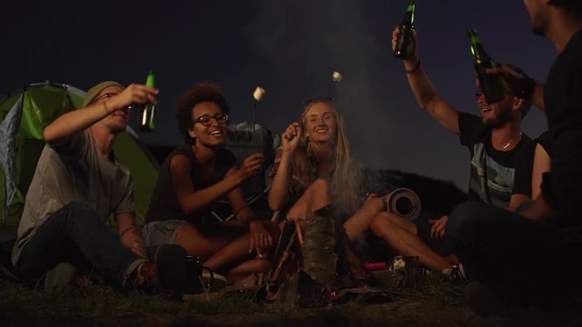 Six friends sitting by fire drinking bottled bear eating fried marshmallow cheering laughing talking smiling. In slowmotion