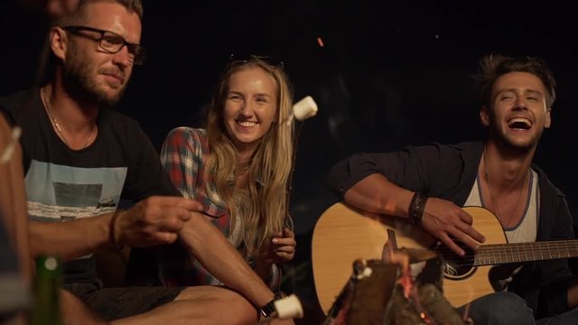 Young Caucasian male playing guitar laughing while friends sitting around listenning talking smiling eating marshmallows. In slowmotion