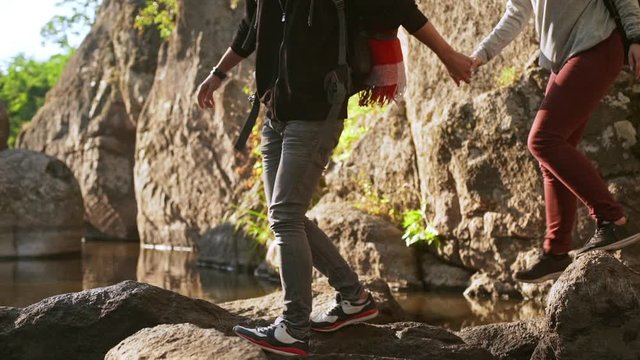 Caucasian male in grey jeans holding hand of female in leggings and carefully walking on rocks near water in slowmotion