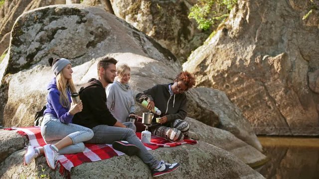 Group of tourists sitting on plaid on rocks near small lake talking drinking from thermoses in slowmotion