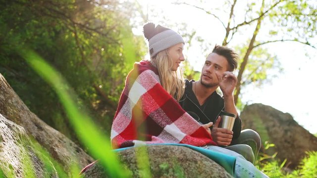 Shy blonde girl in hat with pompon covered in plaid sitting outdoors with handosme male removing eyelash from her face. In slowmotion