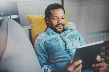 Closeup view of young bearded African man spending chill time in sofa and looking at tablet modern apartment.Concept  people enjoying mobile devices.Blurred background.