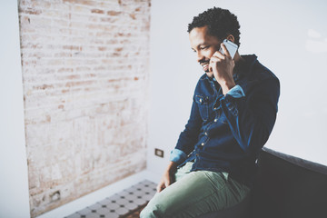 Closeup of young bearded African man making conversation with friends on smartphone at modern home office.Concept people using mobile devices.Blurred wall background.