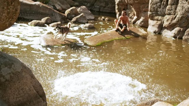 Topless Caucasian male with tattoes jumping from rock to river while another man looking playing with husky dog in slowmotion