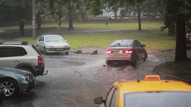 Rain pouring down on pavement, a taxi and cars turns into flood. Another car passes by, water under it splashes around.
