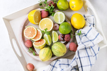 Variety of whole and sliced citrus fruits pink tiger lemon, lemon, lime, mint and exotic lichee on white tray with kitchen towel over white concrete textured background. Top view. Healthy eating