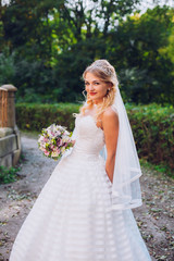 Fototapeta na wymiar Beauty bride in bridal gown with bouquet and lace veil on the nature. Beautiful model girl in a white wedding dress. Female portrait in the park. Woman with hairstyle. Cute lady outdoors