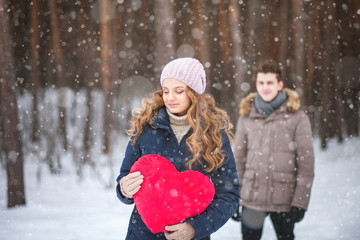 Girl standing and holding a big heart, the guy behind her in the winter forest - sadness, love, expectation, gift, surprise, Valentine's Day